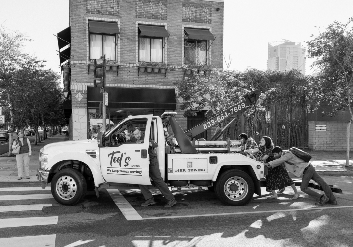 A group of people pushing a broken down tow truck that has “Ted’s Towing” written on the driver’s side door